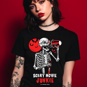 Pode incluir: Camiseta preta com um desenho de esqueleto usando &oacute;culos 3D e segurando um megafone vermelho. Uma lua vermelha e morcegos no fundo. O texto na camiseta diz "SCARY MOVIE JUNKIE" e "MY HAUNTED SPACE".
