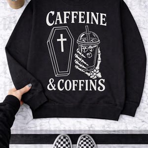 May include: Black sweatshirt with the text "CAFFEINE & COFFINS" in white. The design includes a coffin with a cross and a skeleton hand holding an iced coffee. A Starbucks cup is visible in the background.