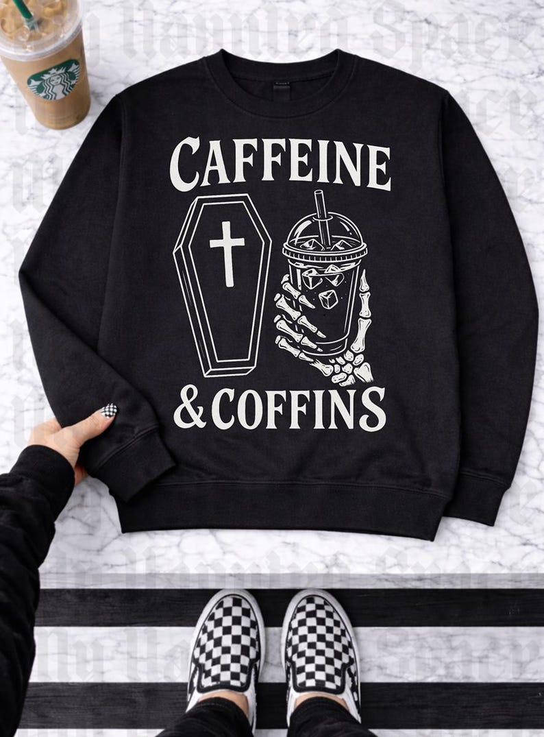 May include: Black sweatshirt with the text "CAFFEINE & COFFINS" in white. The design includes a coffin with a cross and a skeleton hand holding an iced coffee. A Starbucks cup is visible in the background.