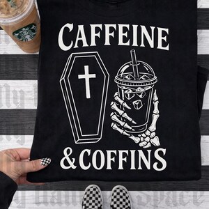 May include: Black t-shirt with the words "CAFFEINE & COFFINS" in white. The graphic includes a coffin and a skeleton hand holding an iced coffee. A Starbucks coffee cup is visible in the upper left corner.