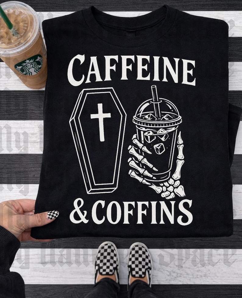 May include: Black t-shirt with the words "CAFFEINE & COFFINS" in white. The graphic includes a coffin and a skeleton hand holding an iced coffee. A Starbucks coffee cup is visible in the upper left corner.
