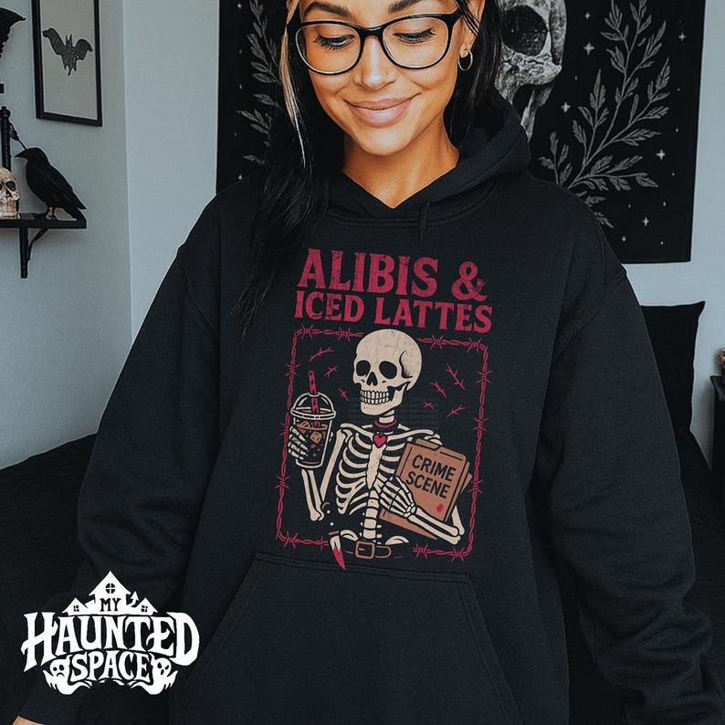 May include: Black hooded sweatshirt with a graphic featuring a skeleton holding a coffee and a book labeled "Crime Scene." The text reads "ALIBIS & ICED LATTES" in pink. The sweatshirt has a front pocket and a "My Haunted Space" logo.