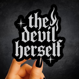 May include: A die-cut sticker featuring the text "the devil herself" in a silver, textured, gothic-style font against a solid black background. The design includes two four-pointed star icons and a flame-like flourish on the letter h.