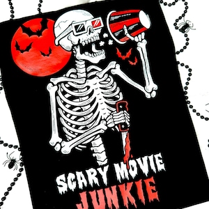 Pode incluir: Camiseta preta com um desenho de esqueleto usando &oacute;culos 3D, segurando um megafone e uma faca ensanguentada. Uma lua vermelha com morcegos ao fundo. O texto diz "Scary Movie Junkie" e "My Haunted Space".