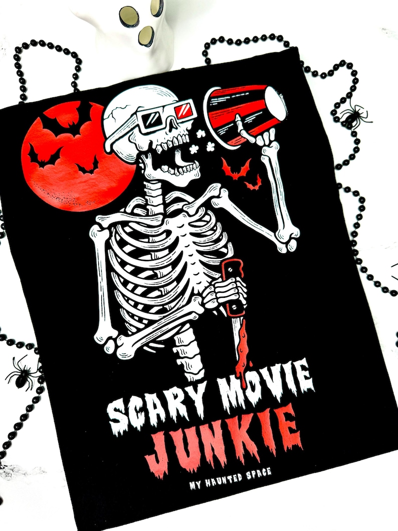 Pode incluir: Camiseta preta com um desenho de esqueleto usando &oacute;culos 3D, segurando um megafone e uma faca ensanguentada. Uma lua vermelha com morcegos ao fundo. O texto diz "Scary Movie Junkie" e "My Haunted Space".