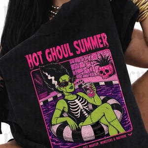 May include: Black t-shirt with a graphic of a green monster in a pool floatie, with the text "HOT GHOUL SUMMER". The monster is wearing sunglasses and a skeleton costume. The image includes the text "NO MELTDOWNS. JUST MAKEUP. MONSTERS & MAYHEM."