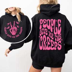 K&ouml;nnte beinhalten: Schwarzer Kapuzenpullover mit einem pinkfarbenen Aufdruck auf der R&uuml;ckseite, der in einer Retro-Schrift "People Give Me The Creeps" lautet. Die Vorderseite des Pullovers hat einen pinkfarbenen Aufdruck einer Skeletthand, die ein Peace-Zeichen macht, mit Flederm&auml;usen, die um sie herum fliegen, und dem Text "People Give Me The Creeps".