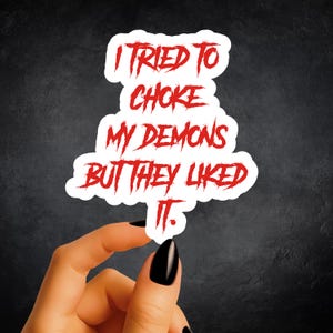 May include: A white die-cut sticker featuring the red text I TRIED TO CHOKE MY DEMONS BUT THEY LIKED IT. The lettering has a jagged, distressed, and scratchy appearance, with a white border surrounding the entire phrase on a dark background.