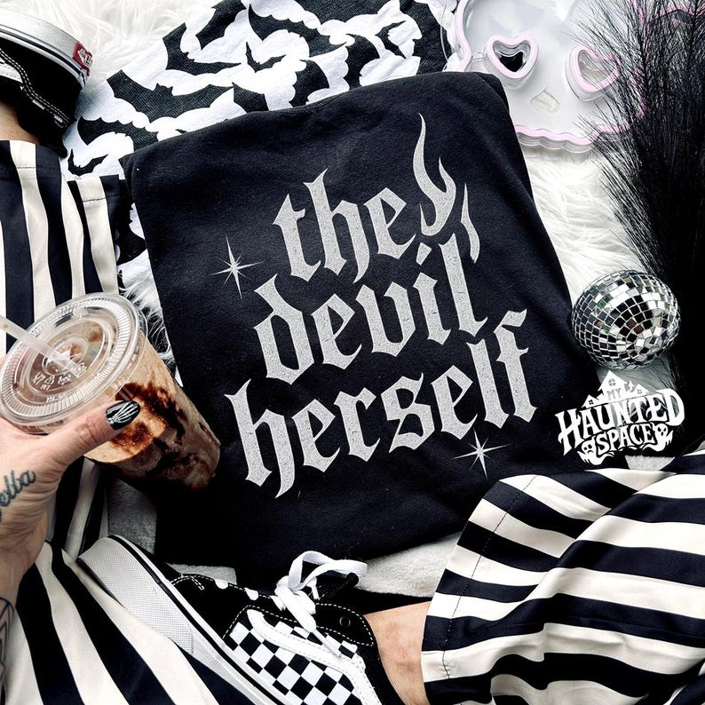 May include: Black hoodie with the text "the devil herself" in white gothic lettering. The image includes a pair of black and white checkered sneakers, a striped tote bag, and a disco ball. A clear plastic cup with a beverage is also visible.