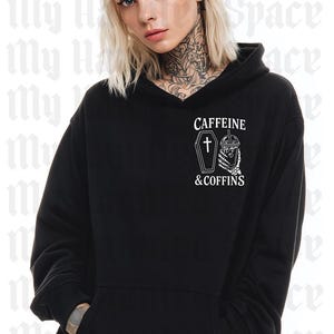 May include: Black hoodie with a front pocket and a hood. The hoodie features the text "CAFFEINE & COFFINS" with a coffin and a coffee cup graphic. The model is wearing the hoodie.