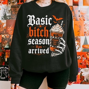 May include: A black long-sleeve sweatshirt featuring a graphic print with the text "Basic bitch season Has arrived" in white and orange gothic lettering. The design includes a skeleton hand holding a pumpkin spice latte and orange bats.