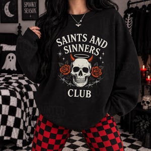 May include: A black long-sleeve sweatshirt featuring a graphic print of a skull with red horns and a halo. The text reads SAINTS AND SINNERS CLUB, surrounded by red roses and barbed wire. The design is centered on the chest of the garment.