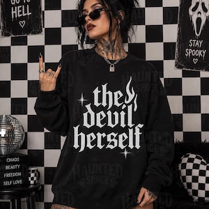Puede incluir: Una sudadera negra de manga larga y cuello redondo con el texto blanco 'the devil herself' impreso en la parte delantera en una fuente gótica estilizada, acentuada con pequeños símbolos de estrellas y un gráfico de llama sobre la palabra 'the'.