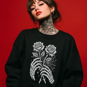 Puede incluir: Sudadera negra con un gráfico blanco de dos rosas sostenidas por manos de esqueleto, con un diseño de telaraña. Las rosas tienen un efecto de goteo. La sudadera es de cuello redondo.