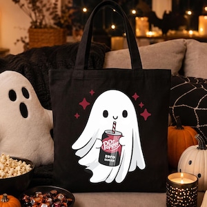 May include: A black tote bag featuring a white cartoon ghost illustration holding a can of Dr Pepper Zero Sugar with a straw. The ghost is surrounded by small red star-shaped accents. The design is centered on the front of the fabric bag.