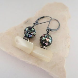 May include: A pair of dangle earrings featuring a dark, mosaic-patterned bead and a rectangular, pearlescent charm. The earrings have a silver-tone leverback closure and are adorned with small, clear crystals. The earrings are displayed on a white surface.