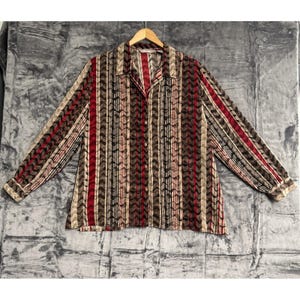 May include: A long-sleeved button-down shirt with a vertical striped pattern in shades of red, brown, black, and beige. The shirt has a collared neckline and buttoned cuffs. The shirt is hanging on a wooden hanger.
