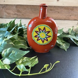 May include: A terracotta-colored bottle with a beaded sunburst design. The bottle has a rounded body and a narrow neck. The sunburst design features yellow, red, and blue beads. The bottle has a distressed finish.
