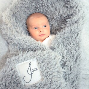 May include: A baby swaddled in a grey fluffy blanket with a white patch that says "Jack" embroidered on it.