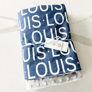 Puede incluir: Una manta para beb&eacute; azul y blanca con el nombre "Louis" repetido en la tela. La manta est&aacute; doblada y atada con una cinta azul. Una peque&ntilde;a etiqueta blanca con la palabra "Pyjamas" est&aacute; unida a la cinta.