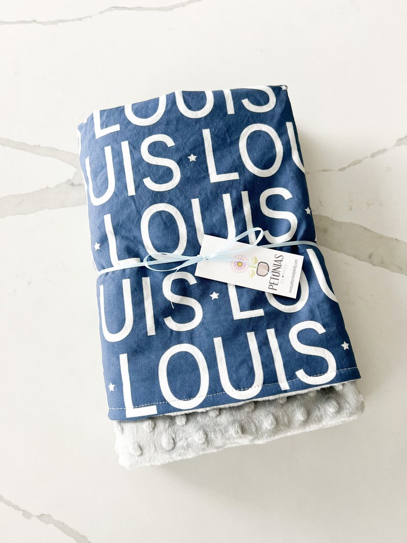 Puede incluir: Una manta para beb&eacute; azul y blanca con el nombre "Louis" repetido en la tela. La manta est&aacute; doblada y atada con una cinta azul. Una peque&ntilde;a etiqueta blanca con la palabra "Pyjamas" est&aacute; unida a la cinta.