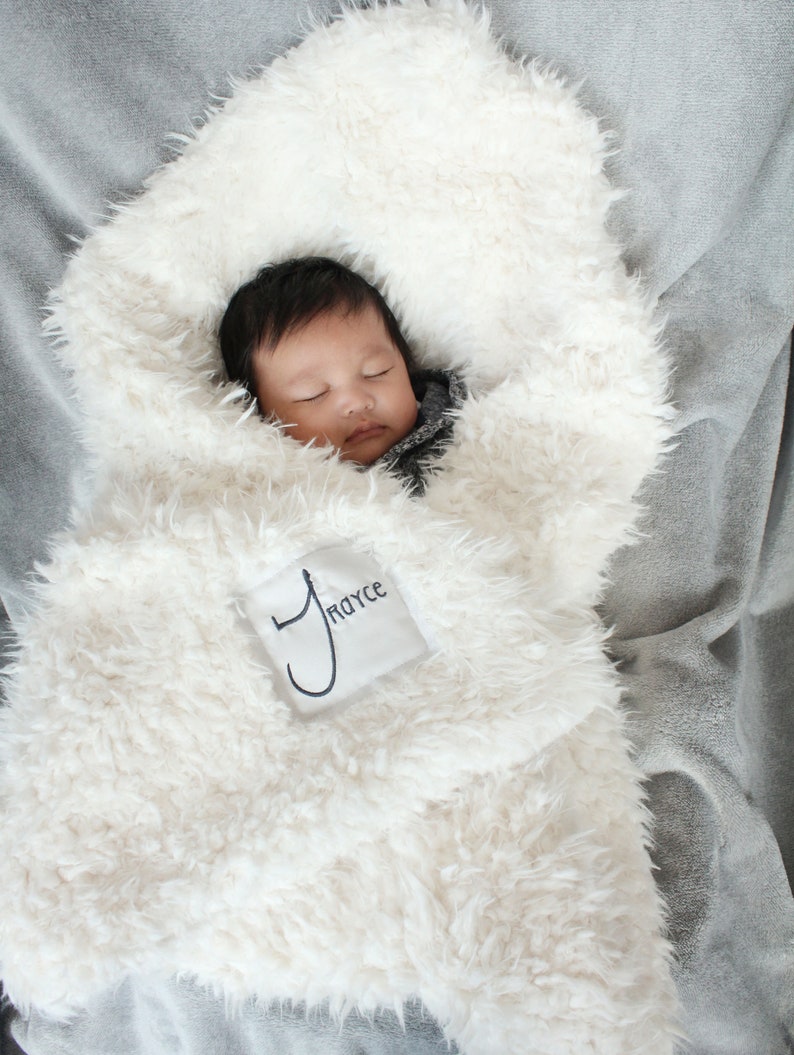 May include: A white faux fur baby blanket with a personalized embroidered name tag that says "Royce". A baby is sleeping peacefully inside the blanket.