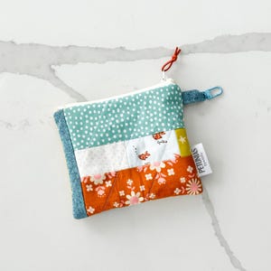 Op de afbeelding: Een klein, gestikt ritsvakje met een patchwork-ontwerp met blauwe, witte, oranje en gele stoffen. Het vakje heeft een wit label met de tekst "PETUNIAS by Ashley" en een blauwe rits.