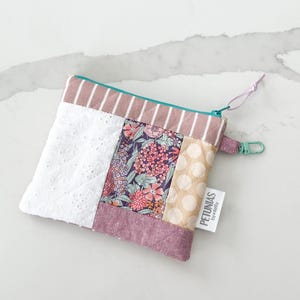 Op de afbeelding: Een klein patchwork-tasje met rits met een bloemenprint, wit kantstof en roze-wit gestreept stof. Het tasje heeft een turquoise rits en een wit label met de tekst "PETUNIAS by Kelly".