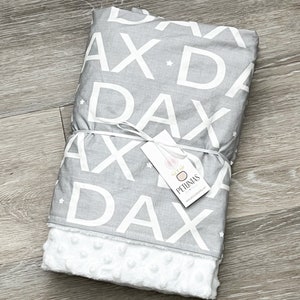 Puede incluir: Una manta para beb&eacute; gris y blanca con un patr&oacute;n repetido de la palabra "Dax" y estrellas. La manta tiene un respaldo de minky blanco suave. La manta est&aacute; atada con una cinta blanca y tiene una peque&ntilde;a etiqueta que dice "Petunias".