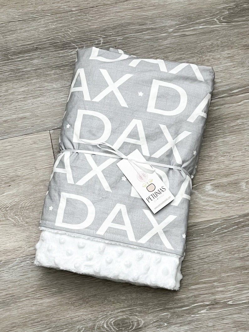 Puede incluir: Una manta para beb&eacute; gris y blanca con un patr&oacute;n repetido de la palabra "Dax" y estrellas. La manta tiene un respaldo de minky blanco suave. La manta est&aacute; atada con una cinta blanca y tiene una peque&ntilde;a etiqueta que dice "Petunias".