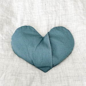 Peut inclure: Un coussin en forme de c&oelig;ur de couleur bleu sarcelle, fait de tissu. Le coussin est pos&eacute; sur une surface textur&eacute;e blanc cass&eacute;. Le coussin a une couture au milieu.