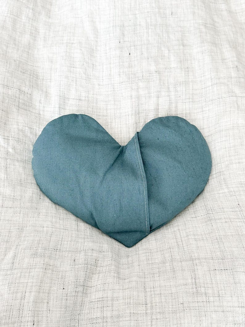 Peut inclure: Un coussin en forme de c&oelig;ur de couleur bleu sarcelle, fait de tissu. Le coussin est pos&eacute; sur une surface textur&eacute;e blanc cass&eacute;. Le coussin a une couture au milieu.