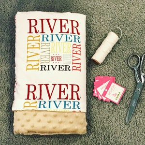 Puede incluir: Una manta de beb&eacute; blanca con el nombre "River" impreso en rojo, amarillo y azul. La manta tiene una textura suave y afelpada, perfecta para acurrucarse.