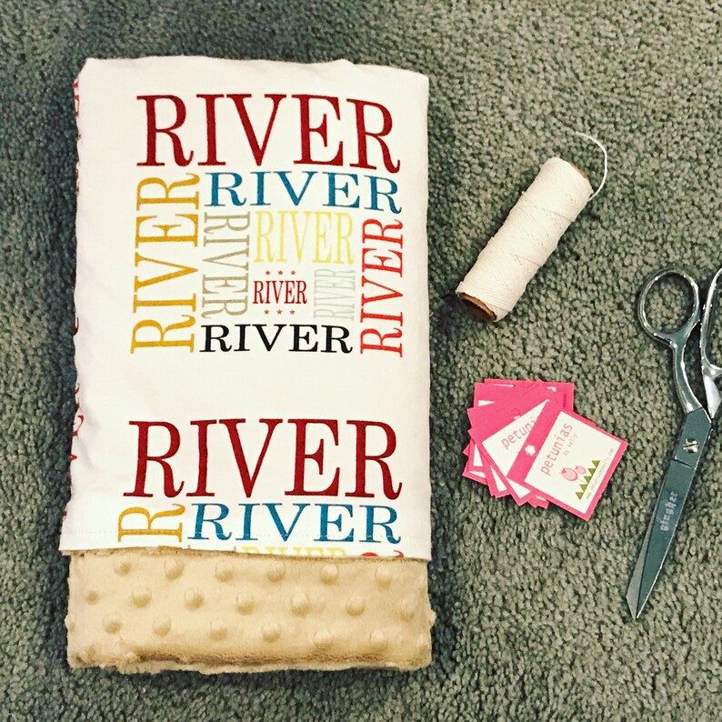 Puede incluir: Una manta de beb&eacute; blanca con el nombre "River" impreso en rojo, amarillo y azul. La manta tiene una textura suave y afelpada, perfecta para acurrucarse.
