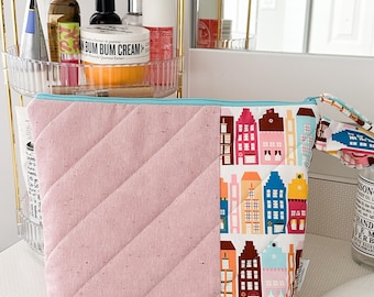 May include: A pink quilted makeup bag with a zipper closure. The front of the bag features a colorful print of houses. The bag is sitting on a white countertop.