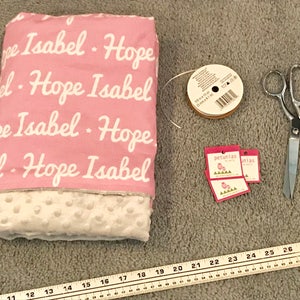 Puede incluir: Una manta de beb&eacute; rosa y blanca con los nombres "Hope" e "Isabel" impresos en ella. La manta est&aacute; rodeada de art&iacute;culos de costura, como tijeras, cinta y etiquetas.