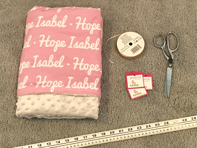 Puede incluir: Una manta de beb&eacute; rosa y blanca con los nombres "Hope" e "Isabel" impresos en ella. La manta est&aacute; rodeada de art&iacute;culos de costura, como tijeras, cinta y etiquetas.