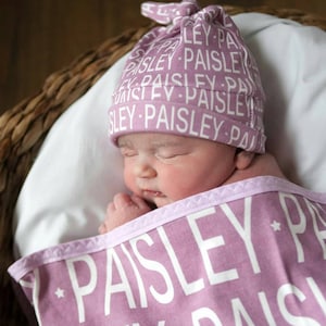 May include: A newborn baby is sleeping in a purple swaddle blanket with the word "Paisley" printed on it. The baby is wearing a purple and white hat with the word "Paisley" printed on it.