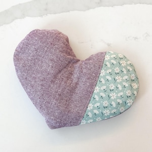Peut inclure: Un objet en forme de c&oelig;ur en tissu lavande et &agrave; imprim&eacute; floral. La partie la plus grande est de couleur lavande unie, tandis que la plus petite section pr&eacute;sente un fond bleu clair avec des motifs floraux blancs. Il s'agit probablement d'un coussin d&eacute;coratif ou d'un sachet.