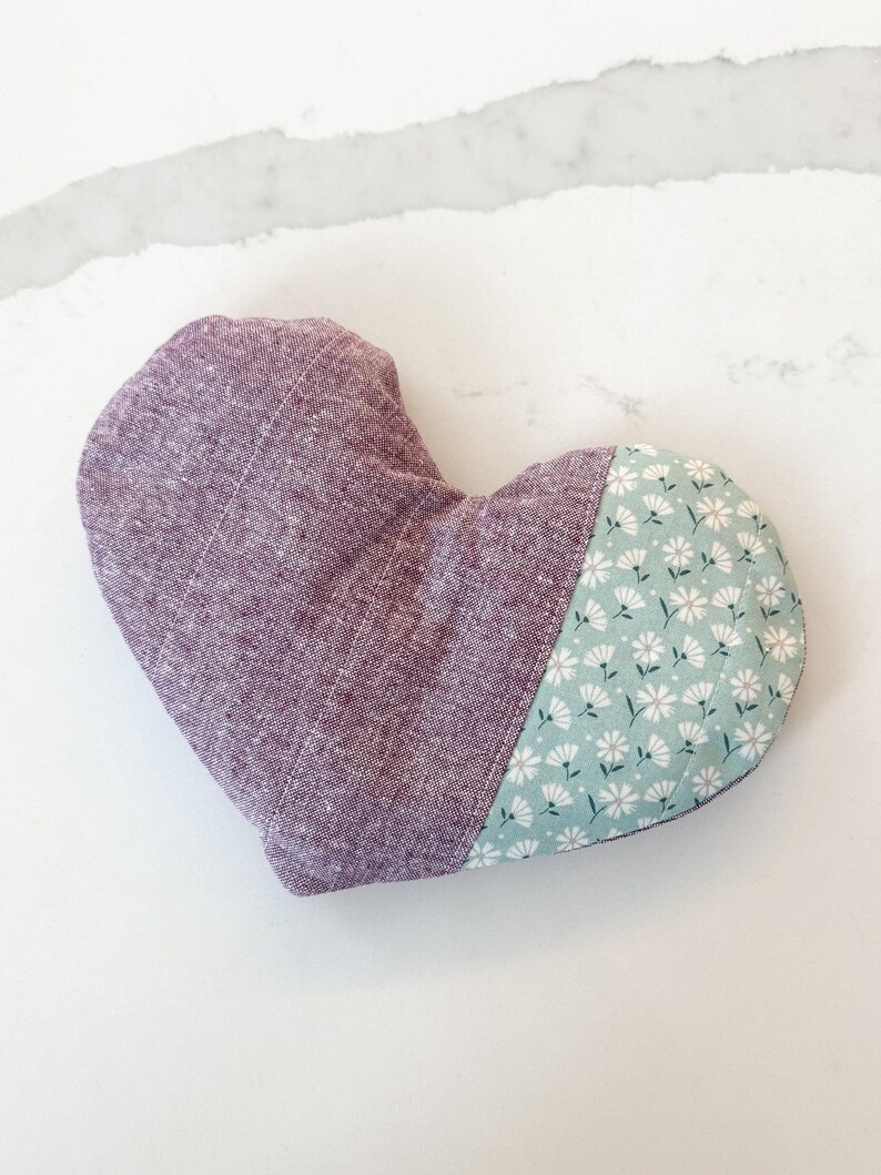 Peut inclure: Un objet en forme de c&oelig;ur en tissu lavande et &agrave; imprim&eacute; floral. La partie la plus grande est de couleur lavande unie, tandis que la plus petite section pr&eacute;sente un fond bleu clair avec des motifs floraux blancs. Il s'agit probablement d'un coussin d&eacute;coratif ou d'un sachet.