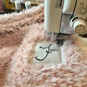 May include: A close-up of a pink fluffy fabric being sewn with a white sewing machine. The fabric has a white label with the name "Fiona" embroidered in black thread.
