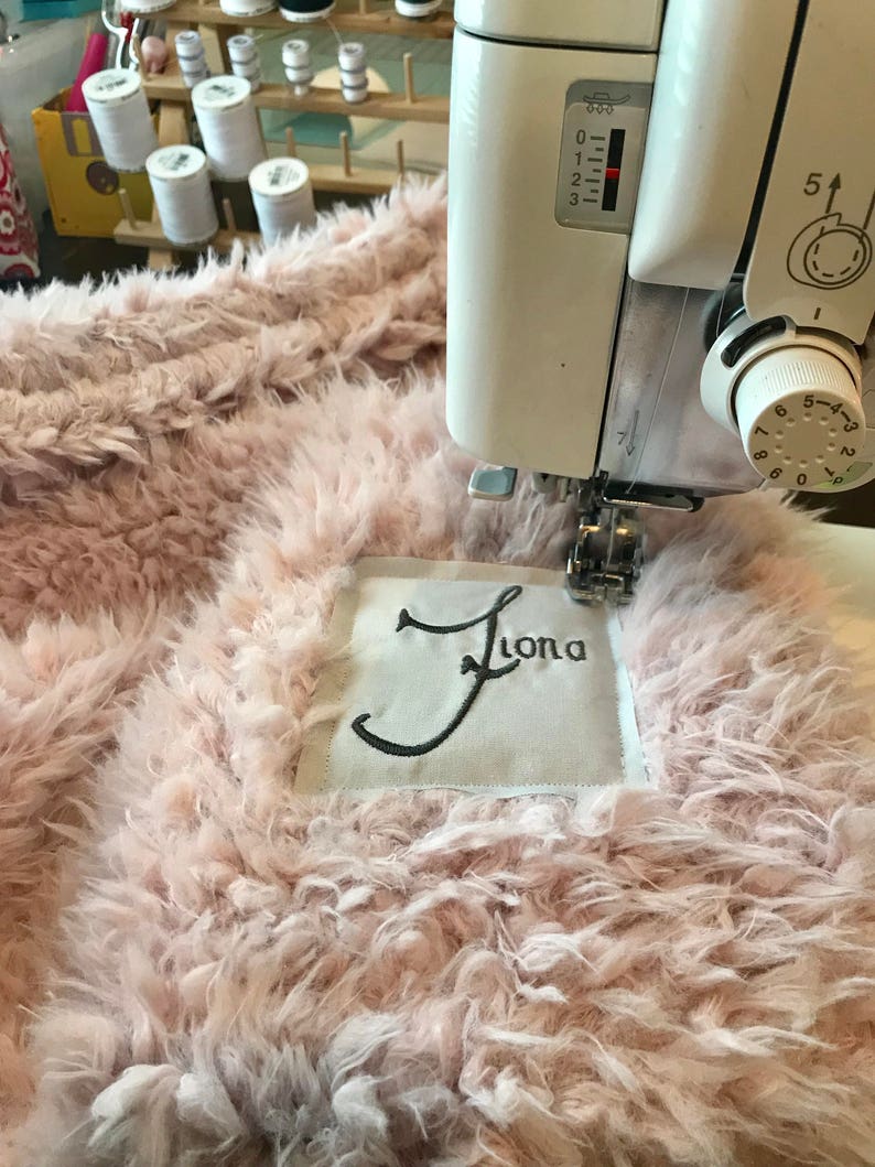 May include: A close-up of a pink fluffy fabric being sewn with a white sewing machine. The fabric has a white label with the name "Fiona" embroidered in black thread.