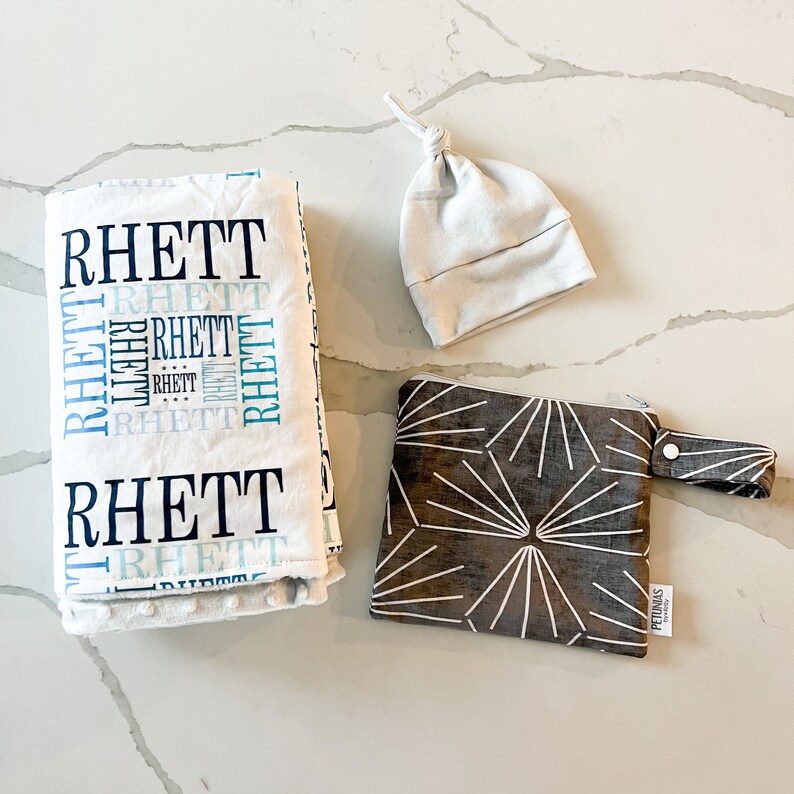 Puede incluir: Una manta de beb&eacute; blanca con el nombre "Rhett" impreso en letras azules y negras. Tambi&eacute;n se incluye un gorro de beb&eacute; de punto blanco y una bolsa con cremallera de estampado geom&eacute;trico marr&oacute;n y blanco.