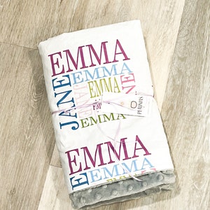 Puede incluir: Una manta de beb&eacute; blanca con el nombre "Emma" impreso en varios colores y fuentes. La manta est&aacute; doblada y atada con una cinta rosa. La manta tiene un lado gris, suave y texturizado.