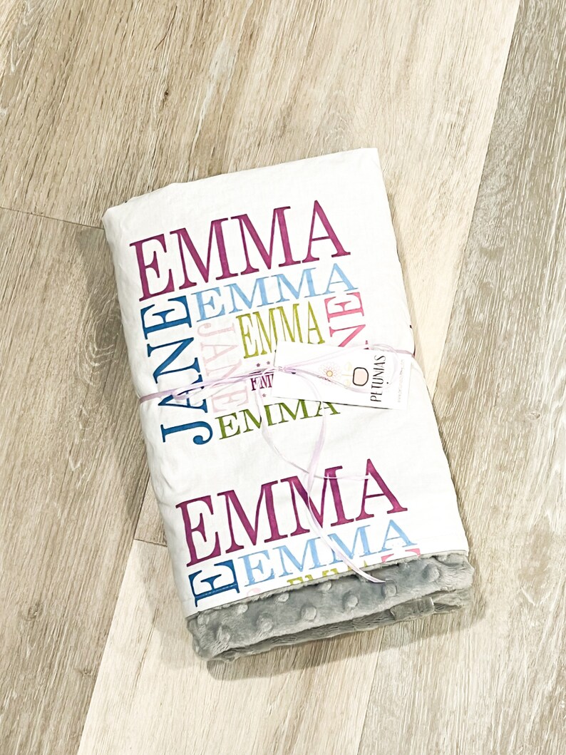 Puede incluir: Una manta de beb&eacute; blanca con el nombre "Emma" impreso en varios colores y fuentes. La manta est&aacute; doblada y atada con una cinta rosa. La manta tiene un lado gris, suave y texturizado.