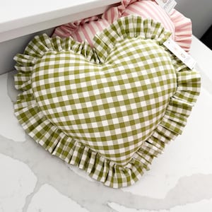 May include: Heart-shaped cushion in a green and white gingham check design, with a ruffled edge. It rests on a white surface. A pink and white striped cushion is partially visible behind it.