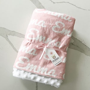 Puede incluir: Manta de beb&eacute; rosa con letras blancas que dicen "Emma" por toda la manta. La manta est&aacute; atada con una cinta blanca y tiene una etiqueta blanca que dice "Petunias by Kelly" y una direcci&oacute;n de sitio web.