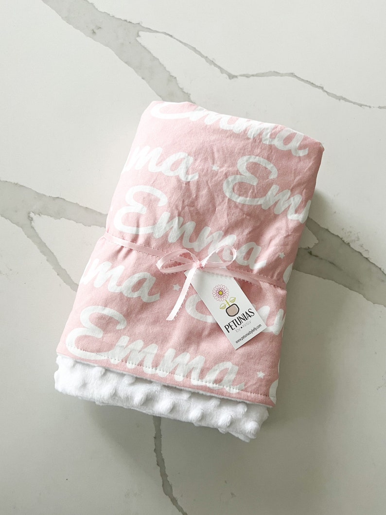 Puede incluir: Manta de beb&eacute; rosa con letras blancas que dicen "Emma" por toda la manta. La manta est&aacute; atada con una cinta blanca y tiene una etiqueta blanca que dice "Petunias by Kelly" y una direcci&oacute;n de sitio web.