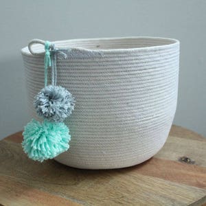 May include: A white rope storage basket with a light gray and mint green pom-pom detail.