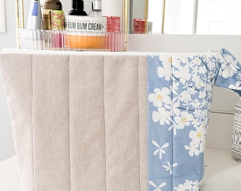May include: A beige quilted cosmetic bag with a blue floral accent. The bag features vertical stitching on the main body and a blue panel with white flowers. The bag is sitting on a white surface.
